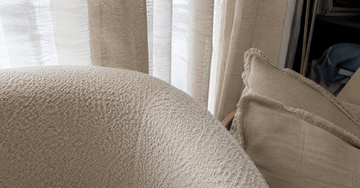 Poltrona in bouclé color crema accanto a cuscini e tende in lino grezzo, trame materiche a confronto