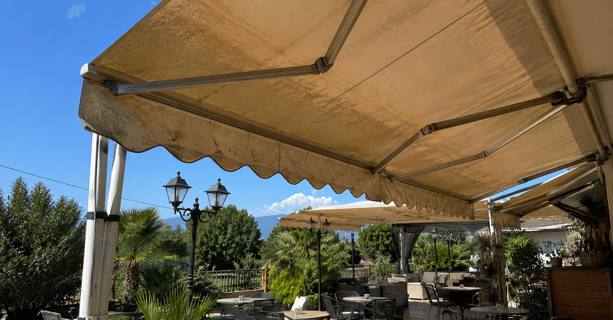 Dehor di un locale con tenda da sole in acrilico dai colori vivi e saturi, tavolini all'ombra sotto il cielo estivo