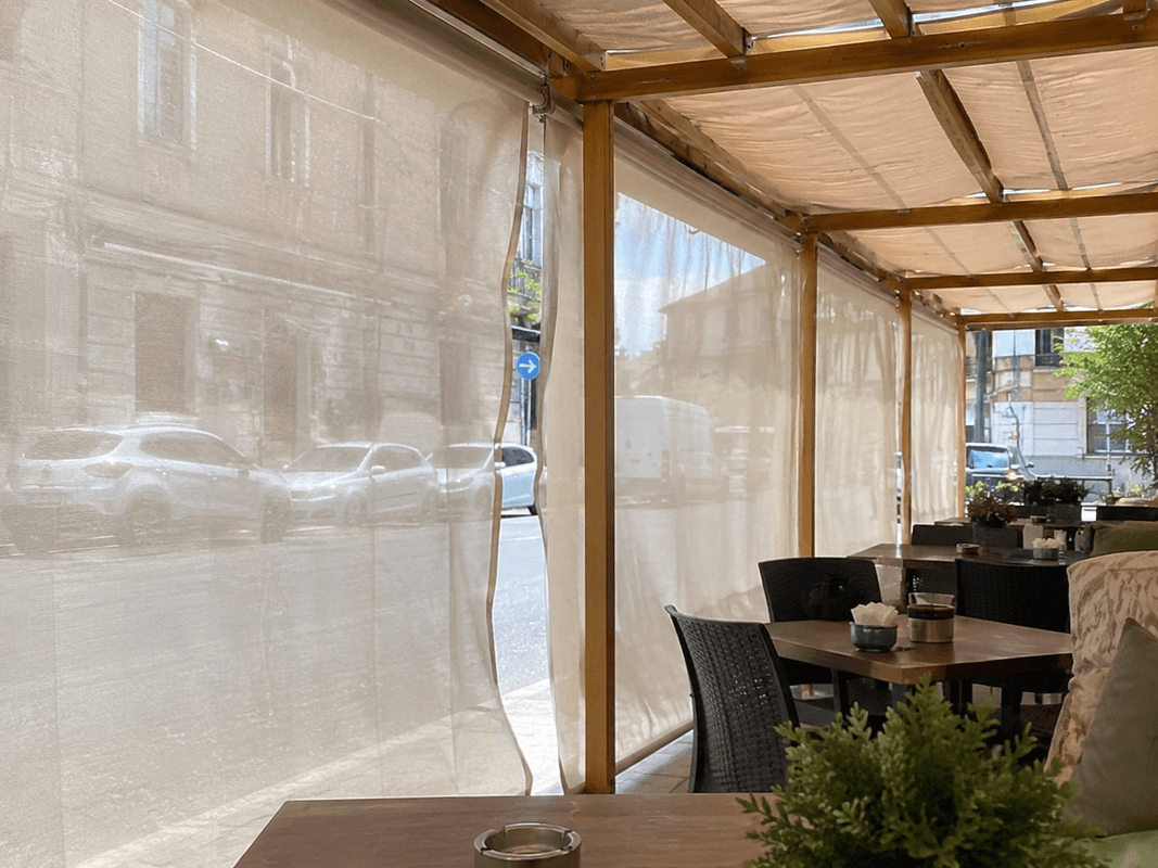 Dehor di un bar con tende divisorie laterali in tessuto chiaro che separano i tavoli dalla strada