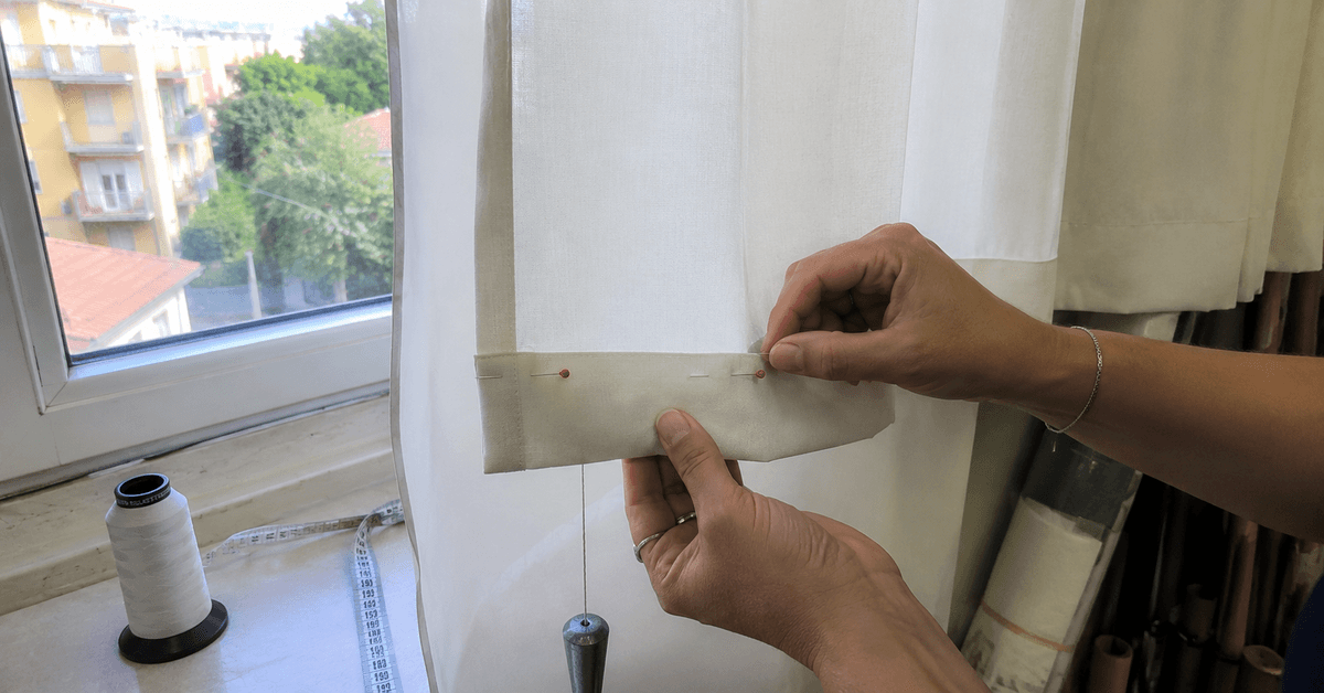 Mani che segnano con gli spilli il nuovo orlo di una tenda chiara appesa alla finestra, con metro a nastro
