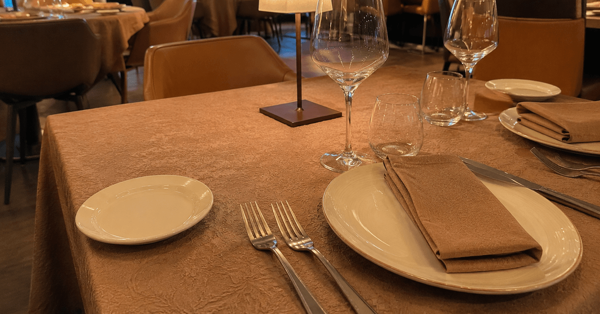 Tavolo di ristorante apparecchiato con tovaglia dai toni caldi tortora con leggera trama jacquard e mise en place elegante