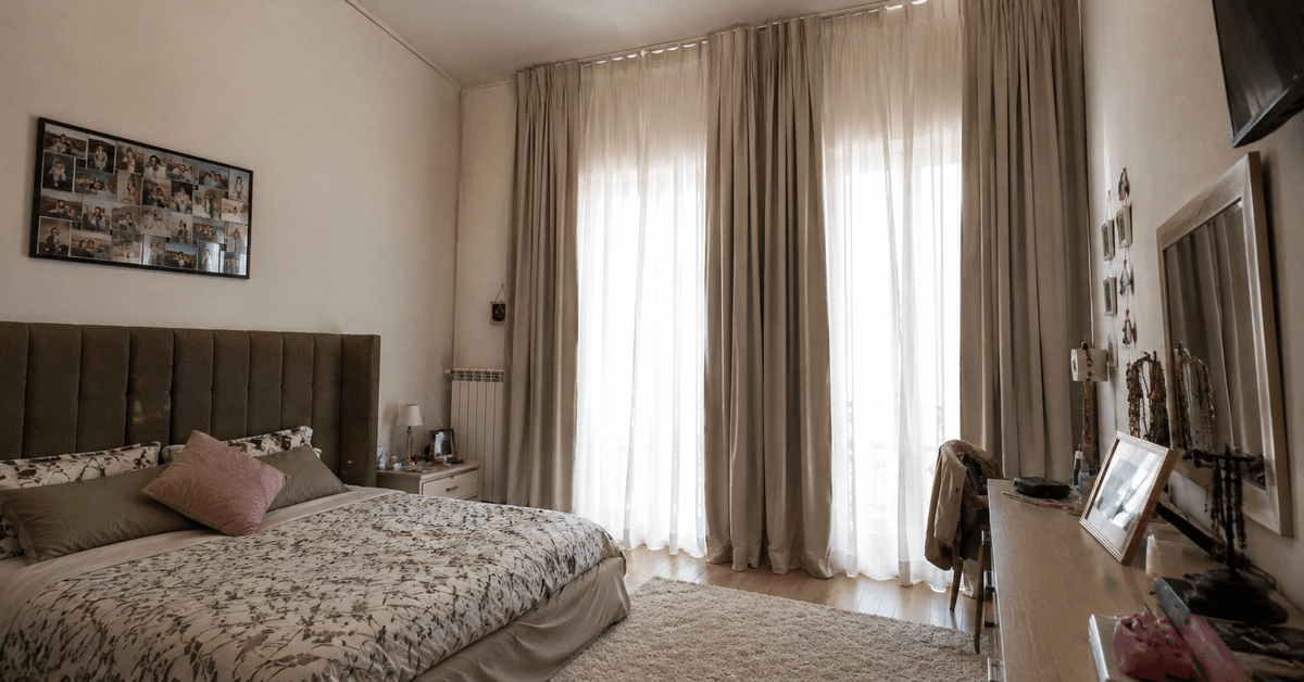 Camera da letto con soffitto alto, tende a tutta altezza abbondanti, testiera imbottita alta e tappeto grande, atmosfera calda e raccolta