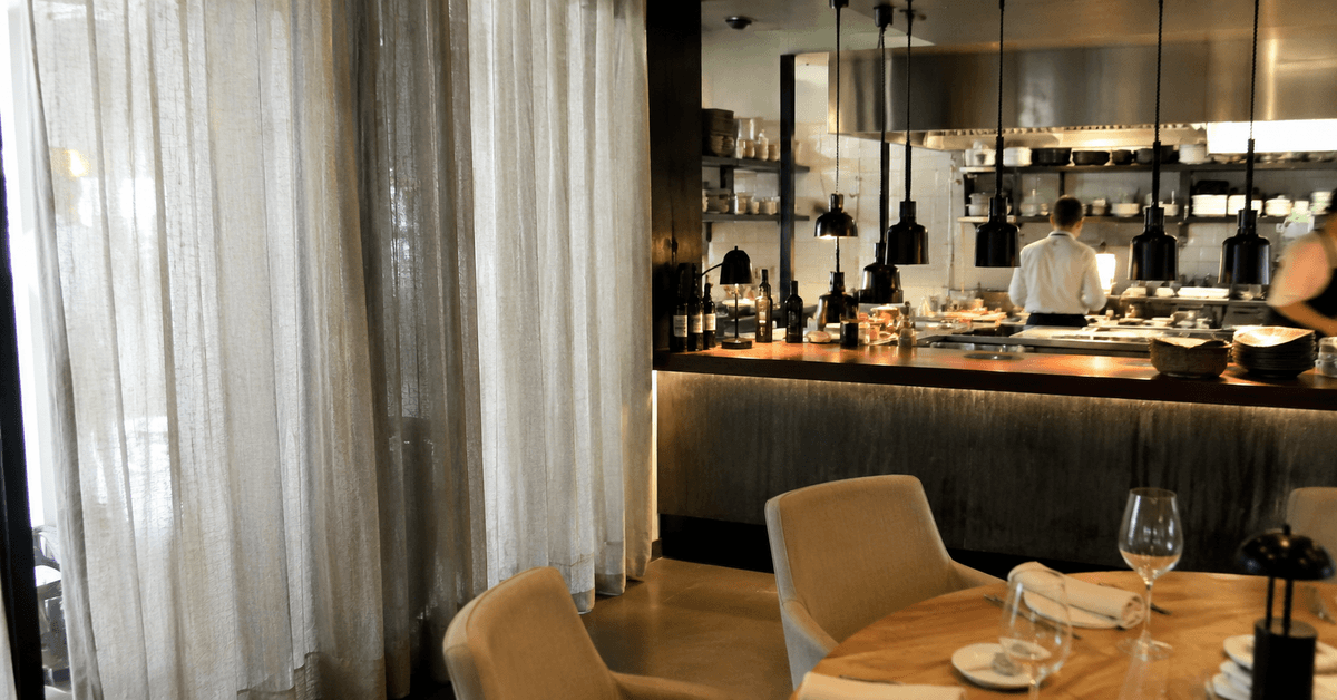 Sala di un ristorante moderno con cucina a vista sullo sfondo e una tenda tecnica leggera vicino al pass
