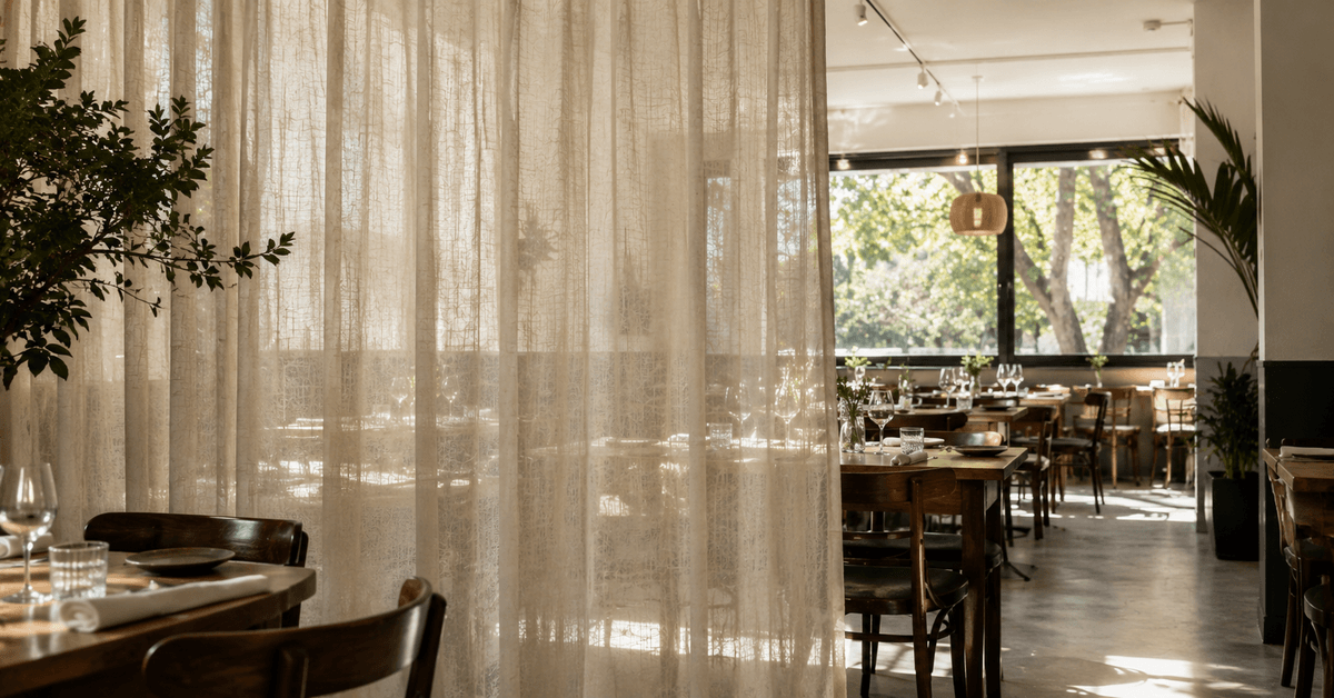 Interno di ristorante con una tenda separ&eacute; leggera che divide due zone tavoli in un ambiente luminoso