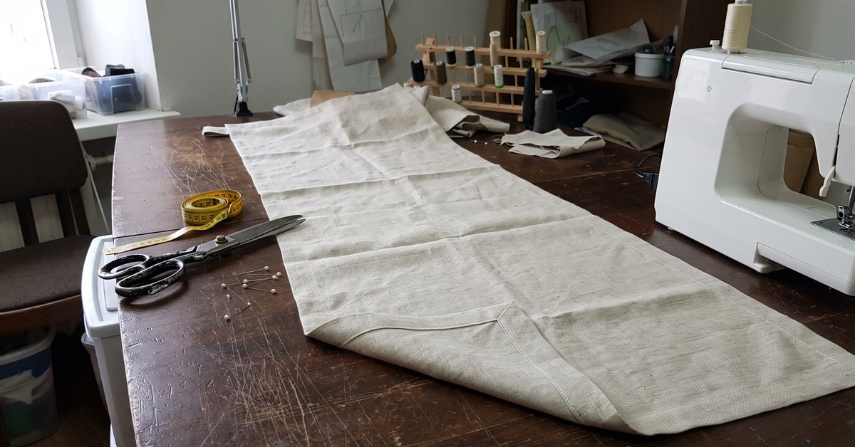 Runner da tavola in lino chiaro cucito su misura, steso sul tavolo del laboratorio con metro, forbici e spilli, angolo rifinito a 45 gradi in primo piano
