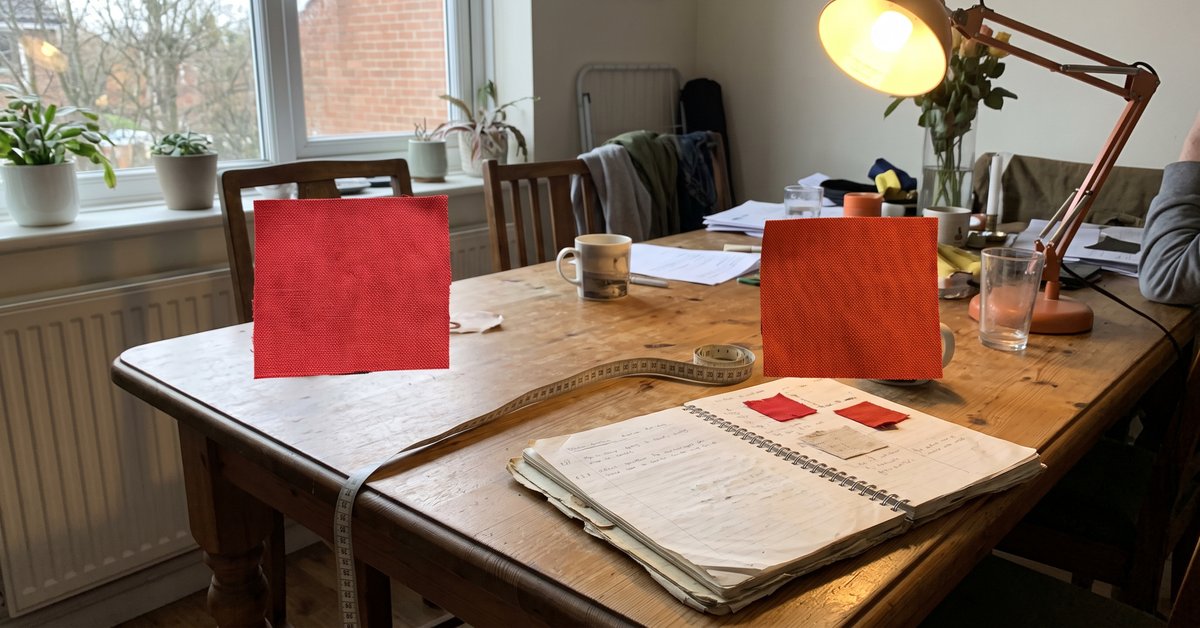 Due campioni dello stesso tessuto rosso a confronto, uno alla luce naturale della finestra e uno sotto una lampada calda, con tonalità diverse