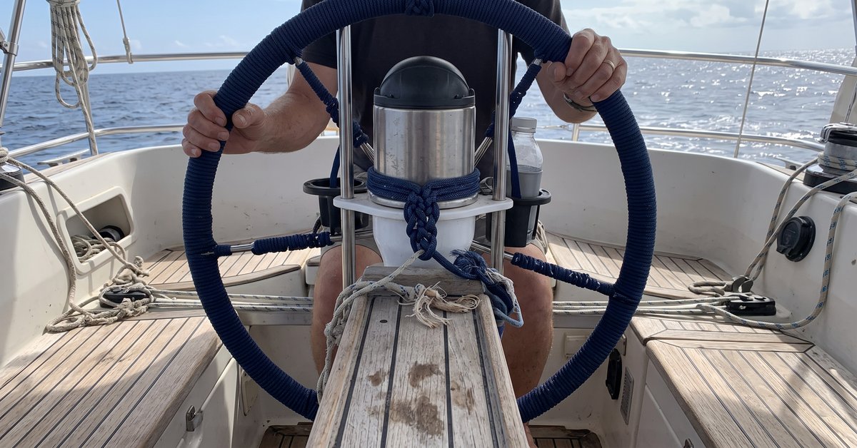 Corona del volante del timone di una barca a vela rivestita in cordino di poliestere cucito a mano, mani che la impugnano, pozzetto in teak sullo sfondo