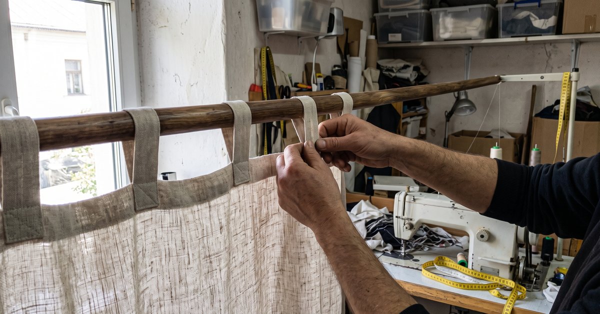 Parte alta di una tenda in lino chiaro con i passanti di tessuto infilati su un bastone di legno, mani che sistemano un passante in laboratorio