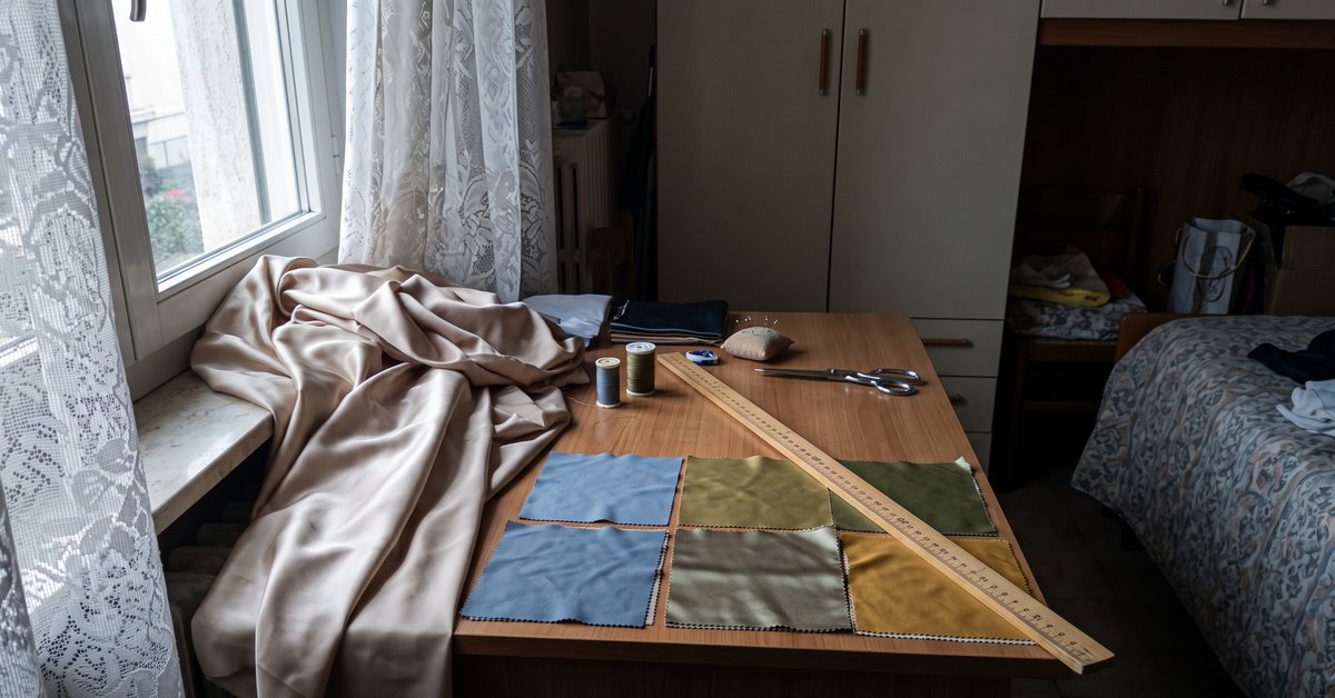 Tende in raso chiaro drappeggiate accanto alla finestra di una camera da letto con riflessi morbidi, e campionature di tessuto lucido e opaco a confronto su un tavolo da lavoro.