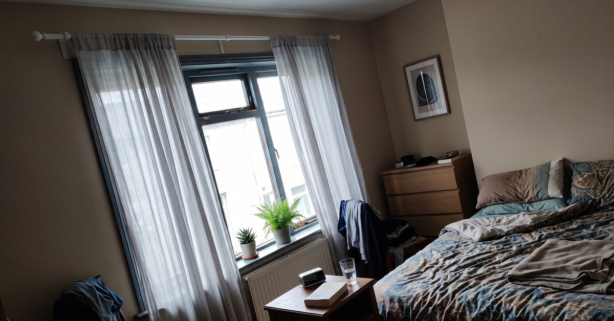 Finestra di una camera esposta a nord con tende chiare e leggere in voile che filtrano una luce soffusa, bastone montato alto e più largo del vetro, letto sullo sfondo