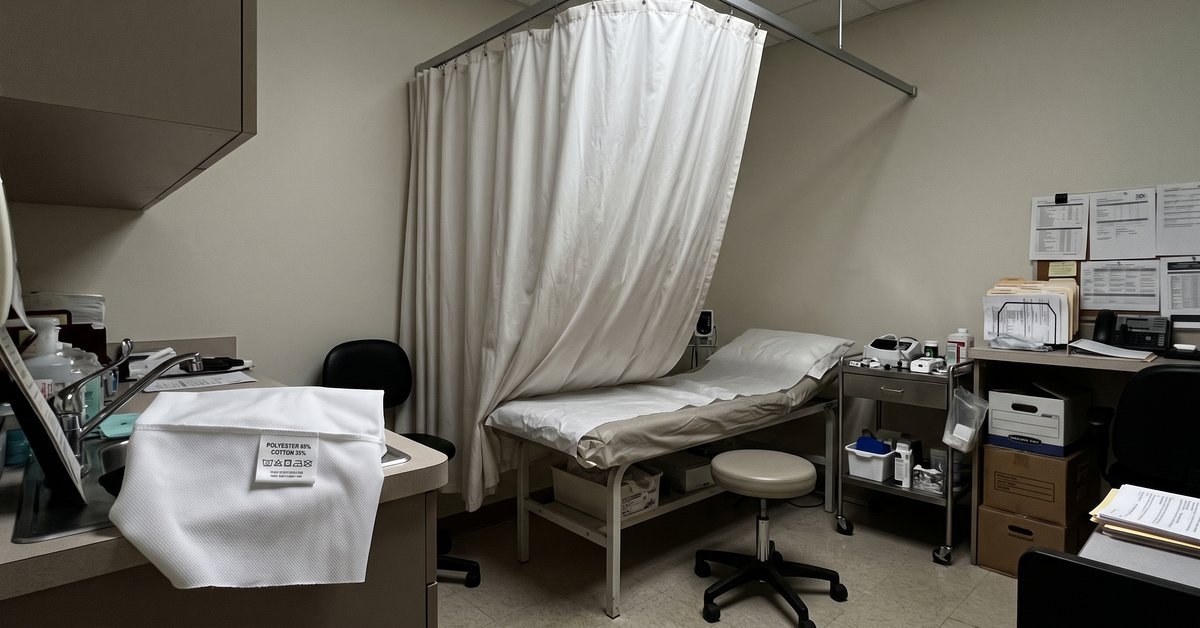 Tenda separatoria chiara su binario a soffitto attorno a un lettino da visita in un ambulatorio, con un campione di tessuto tecnico e la sua etichetta in primo piano