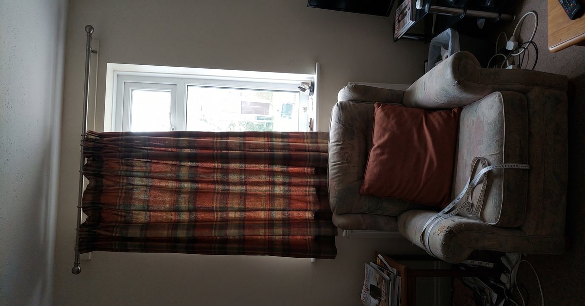Tenda a quadri tartan sui toni caldi di ruggine e verde scuro a una finestra di un soggiorno, con un cuscino terroso su una poltrona e un metro da sarto sul bracciolo