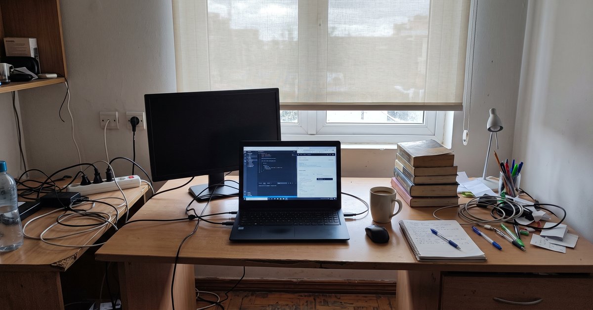 Scrivania con portatile e monitor accanto a una finestra con tenda a rullo chiara filtrante nello studio di casa
