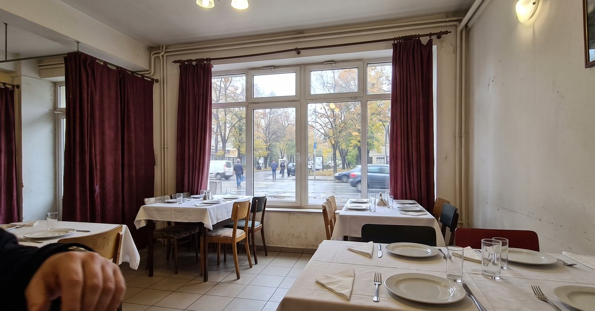 Sala di un ristorante con grande vetrata e tende pesanti scostate ai lati, tavoli apparecchiati vicino al vetro in una giornata autunnale