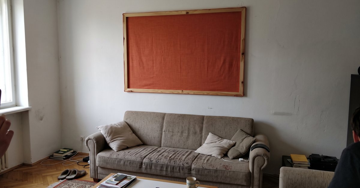 Ampio pannello di tessuto in tinta calda teso su un telaio di legno e appeso a una parete di soggiorno sopra un divano, come alternativa al quadro.